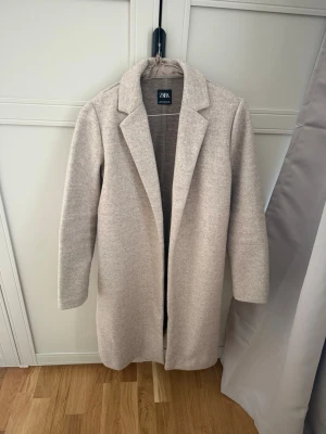 Beige kappa från ZARA med klassisk krage - Stilren beige kappa från ZARA med rak passform och klassisk krage. Kappan har diskreta fickor på sidorna och är tillverkad i ett mjukt, ull-liknande material. Perfekt för dig som vill ha en enkel men snygg look till hösten eller vintern. Använts endast få gånger.
