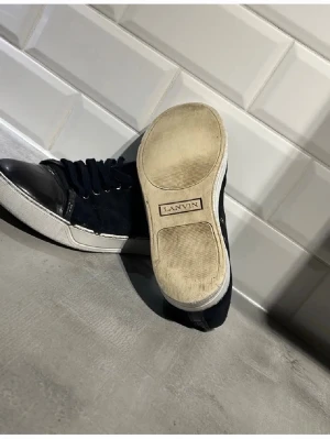 Lanvin svarta låga sneakers - Snygga svarta sneakers från Lanvin med mocka på sidorna och glansiga tåpartier i läder. Klassisk låg modell med vita sulor och svarta skosnören. Perfekta för dig som gillar stilrena och exklusiva sneakers.