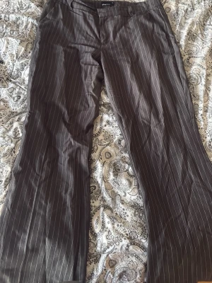 Bruna kostymbyxor med smala ränder - Snygga bruna kostymbyxor med vita smala kritstrecksränder. Byxorna har klassisk rak passform och pressveck framtill. Perfekta för en stilren och dressad look. Stängs med knapp och dragkedja framtill.