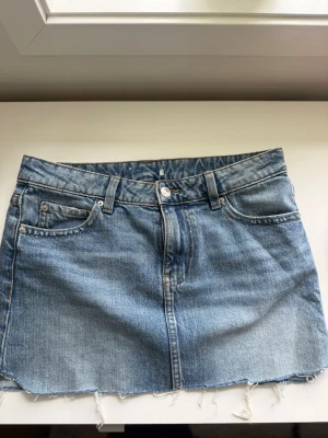 Blå jeanskjol med slitningar - Snygg blå jeanskjol i klassisk modell med fransig kant nedtill. Kjolen har knapp- och dragkedjestängning framtill samt dekorativa bakfickor med lock och knapp. Ursprungligen ifrån Gina Tricot.
