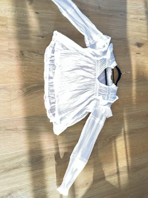 Vit romantisk blus med volangdetaljer - Säljer en luftig vit blus med långa ärmar och volangdetaljer vid axlar och halsringning. Blusen har en lätt transparent känsla och smock vid axelpartiet. Perfekt för en trendig och feminin look.