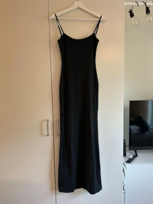 Svart långklänning från Bershka - Säljer en svart långklänning från Bershka med smala axelband och rak passform. Klänningen har en minimalistisk design och är tillverkad i ett mjukt material som känns skönt mot huden. Perfekt för dig som gillar enkel och stilren look.