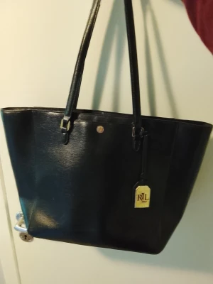 Svart toteväska från Ralph Lauren - Stilren svart toteväska från Ralph Lauren med två långa axelband och guldfärgad RL-detalj framtill. Väskan är tillverkad i slätt konstläder och har en klassisk, rymlig form som passar perfekt för dig som vill ha plats för allt du behöver. Nyskick