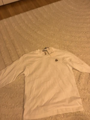 Vit långärmad topp från Moncler - Säljer en stilren vit långärmad topp från Moncler med rund hals och diskret Moncler-logga på bröstet. Tillverkad i mjuk bomull som känns skön mot huden. Perfekt till jeans eller kjol för en clean look.