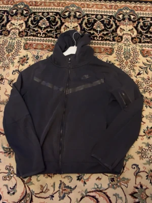 Svart Nike hoodie med dragkedja - Snygg svart hoodie från Nike med hel dragkedja framtill och huva. Diskret Nike-logga på bröstet och en cool detalj med svart band över bröstet. Ficka med dragkedja på ena ärmen. Perfekt för en sportig och avslappnad stil.