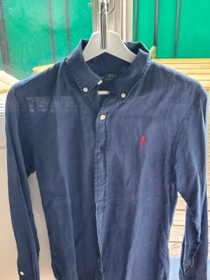 Mörkblå linneskjorta från Polo Ralph Lauren - Klassisk mörkblå skjorta från Polo Ralph Lauren i svalt linne. Skjortan har lång ärm, knappar framtill och ikonisk röd logga broderad på bröstet. Perfekt till jeans eller chinos för en clean och stilren look.
