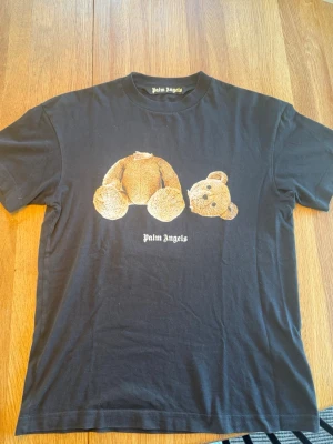 Svart Palm Angels t-shirt med björntryck - Svart t-shirt från Palm Angels med ikoniskt tryck av en nallebjörn utan huvud på framsidan och stor Palm Angels-logga i vitt på ryggen. Klassisk passform och rund hals. Tillverkad i mjuk bomull som känns skön mot huden. Perfekt för dig som gillar streetwear och statement-plagg. Är i storlek s men kan användas till m också. 