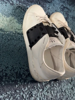 Valentino opens med svarta spikar  - Snygga vita sneakers från Valentino med bred svart läderrem över vristen dekorerad med svarta nitar. Klassisk låg modell med vita snören och diskret logga på plösen. Perfekta för dig som vill ha en edgy och exklusiv look.