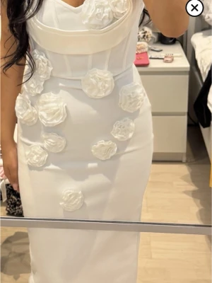 Vit långklänning med blommiga detaljer student kläning - Supersnygg vit långklänning med figurnära passform och stora 3D-blommor framtill. Klänningen är axelbandslös och har en elegant, strukturerad överdel som ger en lyxig känsla. Perfekt för dig som vill sticka ut med en unik och trendig look.