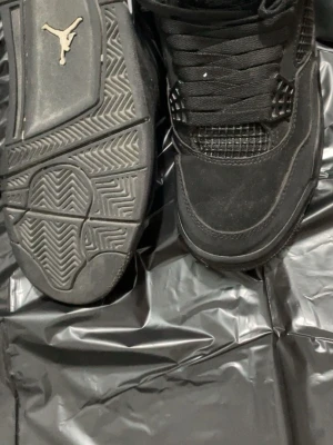 Svarta Air Jordan 4 Retro sneakers - Säljer ett par helsvarta Air Jordan 4 Retro sneakers med klassisk mesh och mockadetaljer. Skorna har synlig Air-enhet i sulan, gummisula med bra grepp och ikonisk Jumpman-logga undertill. Snygg siluett och coola detaljer som gör dem till ett måste för sneakernördar.