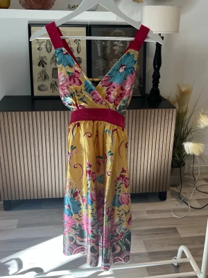 Blommig klänning med omlott - Färgstark klänning med blommigt mönster i rosa, blått och grönt på gul botten från fröken söt. Klänningen har breda axelband, omlott framtill och markerad midja med knytband bak. Tillverkad i ett glansigt material som känns lyxigt och somrigt. Står ingen storlek men skulle säga S. Axelbredd: 26cm. Byst: 32cm. Midja: 36cm. Längd: 93cm.