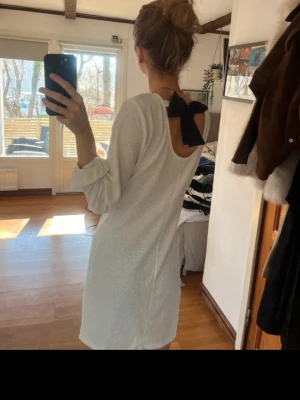 Vit glittrig klänning med rosett - Supersnygg vit kortklänning med glittrigt tyg och lång ärm. Klänningen har öppen rygg med en stor svart rosett som detalj. 
