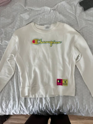 Vit sweatshirt från Champion M - Vit sweatshirt från Champion med broderad logga i gult, blått och rosa på bröstet. Tröjan har rund halsringning, långa ärmar och en patch med Champion-märke och text nertill. Tillverkad i bomull och polyester.