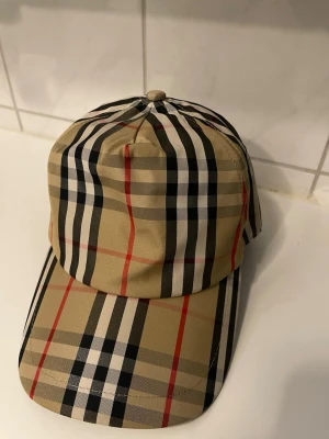 Beige keps med rutigt mönster Burberry - Säljer en klassisk beige keps från Burberry med det ikoniska rutiga mönstret i svart, vitt och rött. Kepsen har böjd skärm och justerbar rem bak. Tillverkad i ett mjukt och lätt material som ger en snygg och tidlös look.