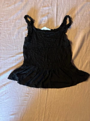 Svart smockad linnetopp med volang - Säljer en svart linnetopp med smockad överdel och breda axelband. Toppen har en volangkant nedtill som ger en söt och trendig look. Perfekt att styla med jeans eller kjol för en chill vibe.