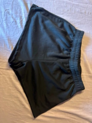 Svarta loose shorts med resårmidja - Säljer ett par svarta shorts med loose passform och bred resår i midjan. Perfekta för chill dagar eller träning. Materialet känns mjukt och stretchigt, troligen polyester eller bomullsmix. Inga synliga loggor eller detaljer.