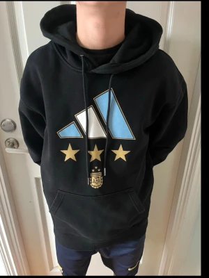 Svart Argentina hoodie från Adidas - Snygg svart hoodie från Adidas med stor Argentina-logga och tre guldstjärnor på bröstet. Klassisk känguruficka framtill, dragsko i huvan och ribbade muddar. Perfekt för dig som älskar fotboll och vill ha en sportig stil. Materialet är mjukt och bekvämt.