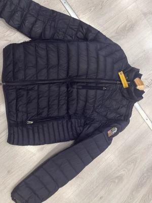 Svart dunjacka från Parajumpers - Snygg svart dunjacka från Parajumpers med gul detalj vid kragen och patch på ärmen. Jackan har två sidofickor med dragkedja och en bröstficka. Lättviktigt material och quiltad design gör den perfekt för kalla dagar. Passar M-L. Pris går diskutera 
