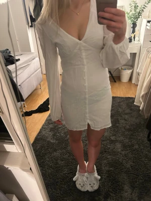 Vit kort klänning med v-ringning - Säljer en vit kort klänning med djup v-ringning och långa, lätt puffade ärmar. Klänningen har knappar framtill och är figurnära med en liten slits nedtill. Perfekt att ha på avslutning. Endast använd en gång 