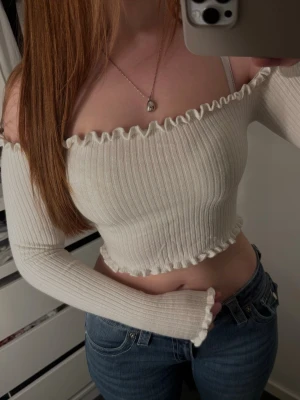 Offshoulder ribbad croppad topp - Supersöt offshoulder topp i ljus ribbad bomull med volangkant upptill och nedtill. Croppad modell som sitter tight och har långa ärmar. Perfekt för dig som vill ha en trendig och bekväm topp till jeans eller kjol.