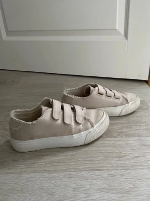 Beige sneakers med kardborreband - Säljer ett par beige sneakers med tre breda kardborreband och råa kanter. Skorna har en vit, tjock sula och är tillverkade i canvasmaterial. Perfekta för en avslappnad och trendig stil. Använda men har mycket kvar att ge. 