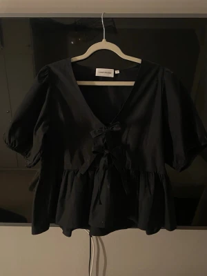 Svart blus med rosett från Carin Wester - Trendig svart blus från Carin Wester med v-ringning och dekorativa rosetter framtill. Blusen har puffiga korta ärmar och en volang nedtill som ger en avslappnad men chic siluett. Perfekt att styla med jeans eller kjol för en cool look.