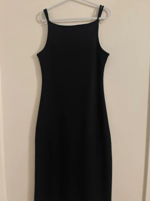 Svart ärmlös långklänning - En stilren svart långklänning med smala axelband och rak halsringning. Klänningen har en figurnära passform och är helt utan mönster eller detaljer, vilket ger en minimalistisk look. Perfekt för dig som gillar enkel och klassisk stil.