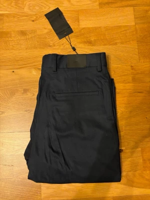 J. Lindeberg Lois Cotton Twill Pant (HELT NYA) - OBS. ALDRIG ANVÄNDA, LAPPEN KVAR.  ORDINARIE PRIS 1800 kr. Snygga marinblå kostymbyxor/chinos från J.Lindeberg i modellen Lois Cotton Twill Pant. Byxorna har klassisk passform, bälteshällor och bakficka med diskret söm. Tillverkade i bomullstwill för en stilren look.