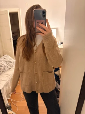 Beige kofta från Zara med fickor - Mysig beige kofta från Zara med v-ringning, guldknappar och två stora fickor framtill. Koftan har en avslappnad passform och ribbade muddar vid ärmslut och nederkant. Perfekt att slänga på sig över en t-shirt för en chill look.