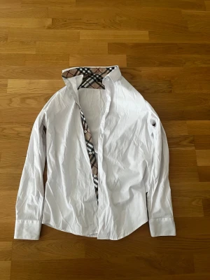 Vit Burberry skjorta  - Säljer en klassisk vit skjorta från Burberry i bomull. Skjortan har långa ärmar och ikoniska beige, svarta och röda rutiga detaljer på insidan av kragen och knappslån. Perfekt för dig som gillar stilrena plagg med exklusiv känsla.