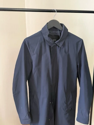Mörkblå car coat från Peak Performance - Snygg mörkblå rock från Stutterheim med minimalistisk design och klassisk krage. Rocken har dold knappslå framtill och två sidofickor. Tillverkad i vattenavvisande material som passar perfekt för regniga dagar.