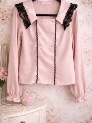 Rosa blus med spetsdetaljer från Honey Cinnamon  - Supersöt ljusrosa blus med långa puffärmar och svarta spetsdetaljer på axlarna. Blusen har dekorativa knappar framtill och svarta spetsband längs bröstet. Materialet ser glansigt ut och ger en elegant känsla. Perfekt för dig som gillar en söt och unik stil.  *Just tried on 