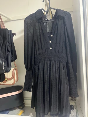 Svart kortklänning med knappar - Snygg svart kortklänning med långärmad design och v-ringning. Klänningen har fina knappar framtill, smock i midjan och ärmarna samt ett diskret prickigt mönster i tyget. Perfekt för dig som gillar en chic och bekväm stil.