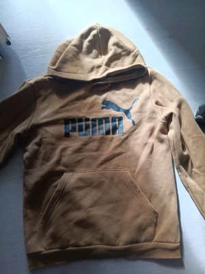 Beige hoodie från Puma med logga - Snygg beige hoodie från Puma med stor svart logga och text på bröstet. Hoodien har en klassisk känguruficka framtill och mjukt, borstat foder på insidan. Perfekt för dig som gillar streetwear och vill ha något bekvämt och stilrent. Materialet är bomullsblandning.