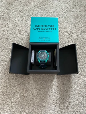 Omega x Swatch MoonSwatch Polar Lights - Snygg Omega x Swatch MoonSwatch Mission on Earth Polar Lights med turkos boett i biokeramik och svart urtavla. Klockan har tydliga vita detaljer, tachymeter och svart textilband med Omega-logga. Perfekt för dig som vill sticka ut med en unik och modern klocka.