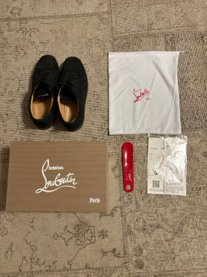 Svarta Christian Louboutin sneakers - Snygga svarta sneakers från Christian Louboutin med klassiska nitar på ovandelen. Skorna har en låg siluett och är tillverkade i mocka med läderdetaljer. Kommer med originalkartong, dustbag och skohorn. Perfekta för dig som vill sticka ut med exklusiv stil.