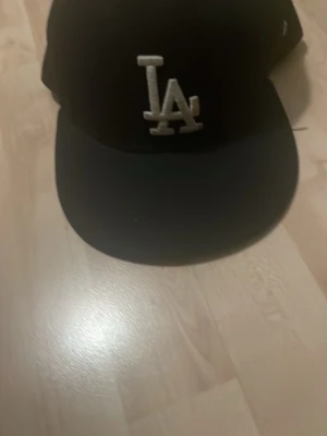 Svart LA-keps från New Era - Säljer en svart keps från New Era med klassisk LA-logga broderad i vitt framtill. Kepsen har rak skärm och justerbar baksida. Perfekt för dig som gillar streetwear och sportig stil.