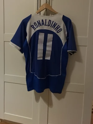 Äkta Blå Brasilien Ronaldinho tröja Nike - Säljer en blå Brasilien fotbollströja från Nike med Ronaldinho och nummer 11 på ryggen. Tröjan har vita detaljer, korta ärmar och Brasilien-emblemet på bröstet. Perfekt för dig som älskar fotboll och vill hylla en legend.