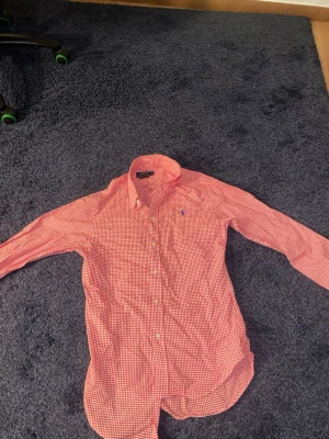 Rutig röd skjorta från Polo Ralph Lauren - Klassisk röd- och vitrutig skjorta från Polo Ralph Lauren med lång ärm och knappar framtill. Skjortan har en liten broderad logga på bröstet och är tillverkad i mjuk bomull. Perfekt för en stilren och avslappnad look.