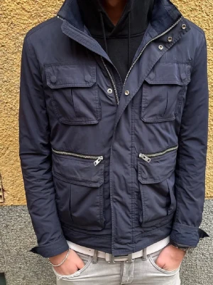 Marinblå vindjacka från J.Lindeberg - Snygg marinblå vindjacka från J.Lindeberg med fyra framfickor med lock och två snedställda dragkedjefickor.Nypris 3500 Jackan har hög krage, tryckknappar och dragkedja framtill samt en diskret logotyp på ärmen. Tillverkad i ett lätt, vindtätt material.