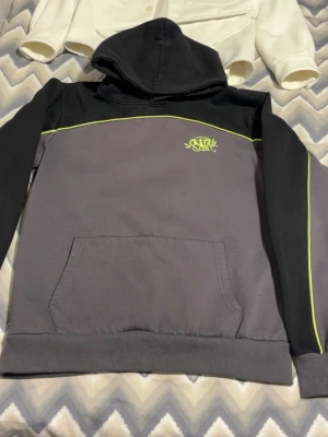 Grå och svart hoodie från Syna World  - Snygg hoodie i grått och svart från SANTA med limegröna detaljer och broderad logga på bröstet. Hoodien har huva, magficka och ribbade muddar. Perfekt för en chill och street-inspirerad stil.