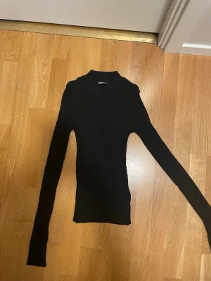 Svart ribbad polotröja - Säljer en svart, ribbstickad polotröja med lång ärm och tight passform. Tröjan har hög hals och är tillverkad i ett stretchigt material som sitter snyggt på kroppen. Perfekt basplagg till garderoben.