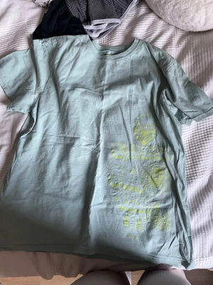 Ljusgrön t-shirt med tryck - Säljer en ljusgrön t-shirt med coolt gult tryck framtill som inkluderar text och tecknade figurer som sol och smiley. T-shirten har rund hals och korta ärmar, perfekt för en avslappnad stil.