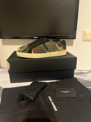 Yves Saint Laurent camo gröna kamouflage skor - Snygga sneakers från Saint Laurent med camo-mönster i grönt, brunt och svart. Skorna har svarta skosnören, beige sula och klassisk låg profil. Perfekta för dig som vill sticka ut med en exklusiv streetstyle-look. Helt nya och i storlek 40 men passar även 41. Kommer med allt og.