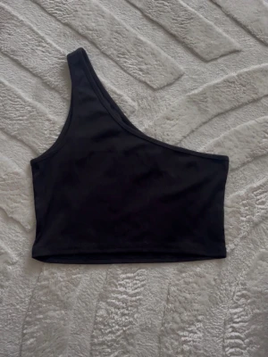 Svart oneshoulder topp - Snygg svart oneshoulder topp i stretchigt material. Perfekt för dig som vill ha en enkel men trendig look. Toppen är ärmlös och har en asymmetrisk axel som ger en cool vibe.