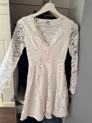 Studentklänning - Säljer en vit kort spetsklänning från bubbleroom, helt slutsåld med v-ringad hals och långa ärmar. Klänningen har ett vackert spetsmönster över hela plagget och en feminin, figurnära passform. Perfekt för sommarens alla tillfällen!