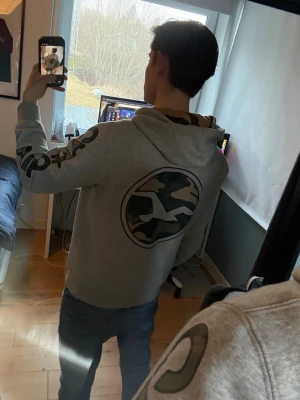 Grå Hollister hoodie med camotryck - Snygg grå hoodie från Hollister med stort camomönstrat logotryck på ryggen och texten 'California' längs båda ärmarna. Hoodien har huva och ribbade muddar. Perfekt för en avslappnad streetstyle-look. Skriv om ni har frågor, priset kan diskuteras.