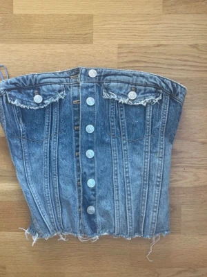 Blå jeanskorsett med knappar - Trendig blå jeanskorsett med fransig nederkant och dekorativa fickor framtill. Toppen har tydliga sömmar, knappar längs hela framsidan och är axelbandslös. Perfekt för en edgy och cool look.