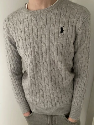 Grå kabelstickad tröja Polo Ralph Lauren - Snygg grå kabelstickad tröja från Polo Ralph Lauren med rund halsringning och den klassiska broderade loggan på bröstet. Tröjan har ribbade muddar vid ärmslut och nederkant, perfekt för en stilren och avslappnad look. Det står storlek M men passar bättre som storlek S. Modellen är 185 och väger 65
