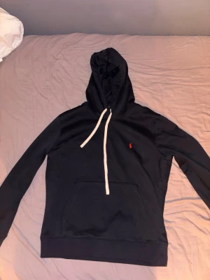 Svart hoodie från Polo Ralph Lauren - Snygg svart hoodie från Polo Ralph Lauren med vit dragsko och klassisk röd logga broderad på bröstet. Hoodien har en stor känguruficka framtill och ribbade muddar vid ärmslut och nederkant. Perfekt för en chill och stilren look.
